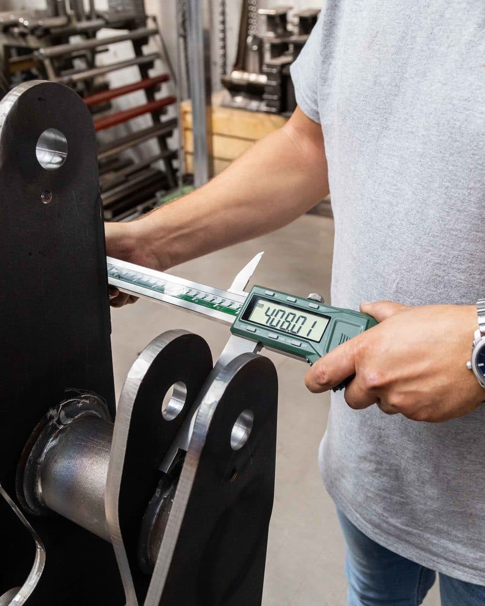 Kapos Atlas employee measuring a machined linkage component with a digital caliper during quality control
